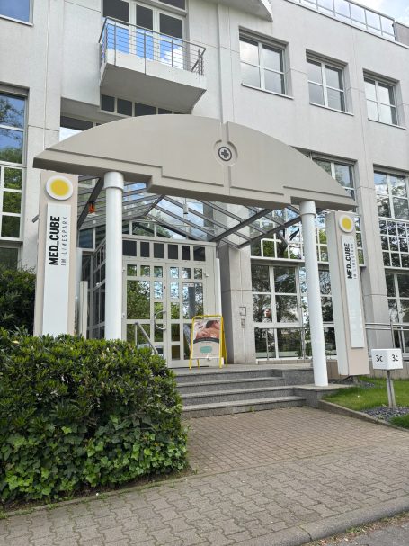 Eingang eines modernen Gebäudes mit Glasfront und Pflanzungen.